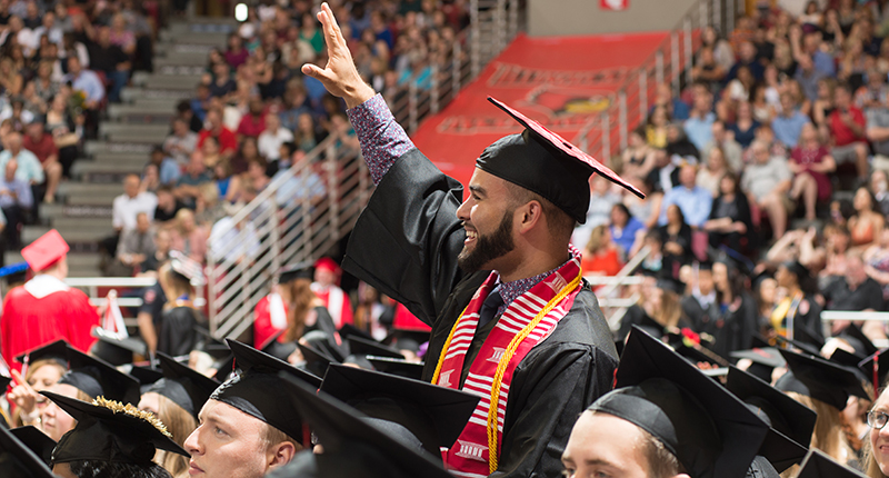 Male in cap and gown standing in arena, waving at crowd.